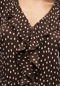 Blouse marron à pois blancs, avec bordure froncée et un seul bouton rond près du col en V.