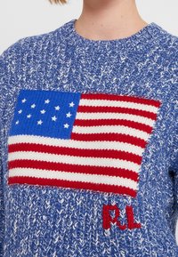Pull en tricot bleu avec un motif texturé. Il présente un patch rectangulaire représentant le drapeau américain en rouge, blanc et bleu, avec les initiales "F.L." brodées en rouge.