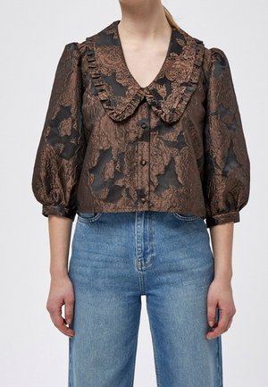 Frau trägt eine schwarze und braune Jacquard-Bluse mit Blumenmuster, Puffärmeln und großem, gerüschtem Kragen, kombiniert mit hoch taillierten blauen Jeans.