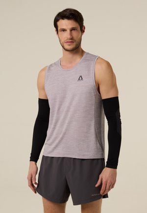 Mann trägt ärmelloses graues Sportshirt, schwarze Armstulpen und dunkelgraue Shorts, steht vor einfarbigem Hintergrund.