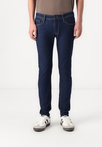 Sötétkék, slim fit farmernadrág, látható varrással, öt zsebből és elülső gombos záródással. Fehér edzőcipővel viselve.