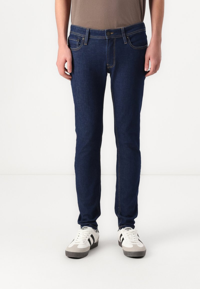 Sötétkék, slim fit farmernadrág, látható varrással, öt zsebből és elülső gombos záródással. Fehér edzőcipővel viselve.