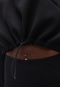 Κοντινό πλάνο της μέσης με μαύρο cropped φούτερ που δείχνει ένα επέχρυσο σκουλαρίκι ομφαλού με καθαρό πολύτιμο λίθο πάνω σε σκούρο δέρμα.