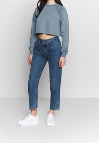 Felpa azzurra corta, jeans in denim a vita alta e sneaker bianchi. La felpa ha maniche lunghe e polsini a coste. I jeans hanno un taglio dritto.
