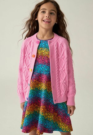 Niña joven con un vestido floral colorido con rayas horizontales de arcoíris y un cárdigan rosa de punto con botones multicolores.