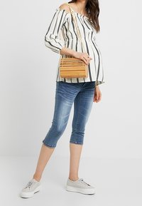 Kompakt trähandväska i ljus tan med horisontella ribbor, bärs kors och tvärs. Modell i randig off-shoulder-top och blå capribyxor, grå sneakers.