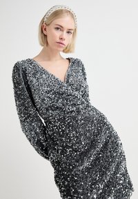 Vestido prateado com lantejoulas, mangas longas, decote em V e design franzido, exibindo uma textura suave e detalhes cintilantes.