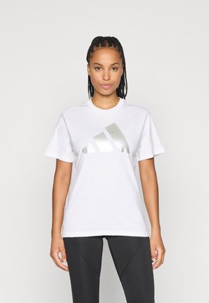 Hvit bomulls T-skjorte med et stort metallisk sølv Adidas-logo på fronten, korte ermer og en avslappet passform. Sammen med svarte tights.
