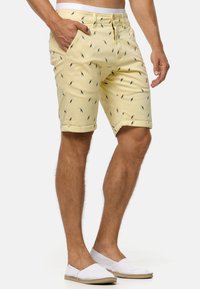 Shorts jaunes avec un imprimé d'oiseaux répétitif, dotés de poches latérales, d'une fermeture éclair et d'un ourlet roulé. Associés à des chaussures blanches à enfiler.
