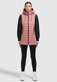 Rosa pufferväst med hög krage och huva, med dragkedja framtill, två sidfickor och quiltad textur. Bärs över en svart långärmad topp.