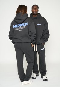 Twee modellen dragen bijpassende donkergrijze hoodies en joggingsbroeken. De hoodies hebben blauwe logo's op de achterkant en op de borst. De broeken hebben zakken en koordjes.