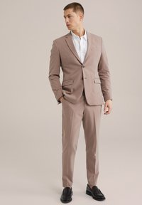 Világosbarna öltöny, testre szabott fazonnal, egy gombos záródással, két elülső zsebbel és passzoló nadrággal. Fehér inggel és fekete cipőkkel viselik.