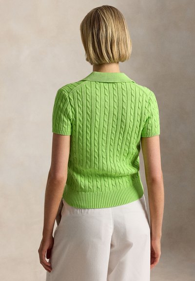 Lyhytukkainen vaaleahiuksinen nainen pitää päällään kirkkaan vihreää, kaapelisukkaa muistuttavaa lyhythihaista villapaitaa ja luonnonvalkoisia housuja, kuvattuna takaa neutraalia taustaa vasten.