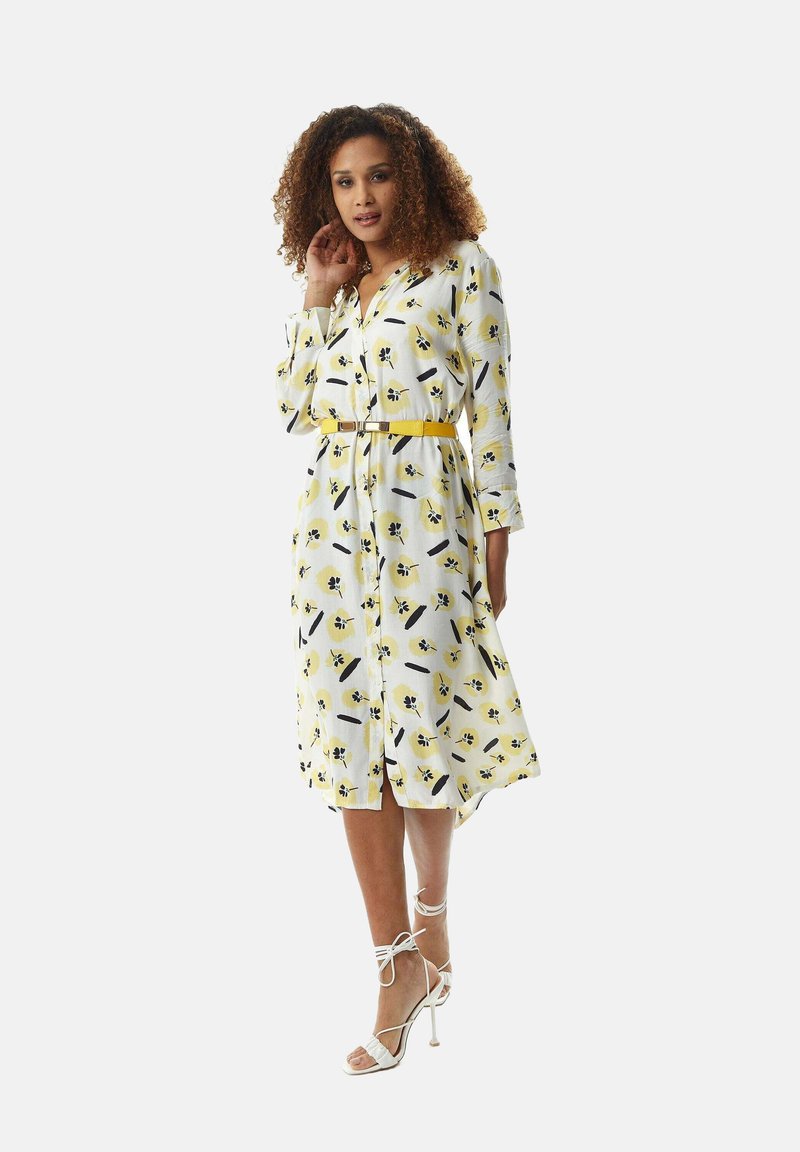 White dress with yellow and black floral print, long sleeves, button front, and belted waist. Paired with white strappy heels.