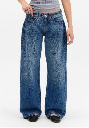 Personne portant un jean denim bleu large et des chaussures noires avec un haut gris court, debout avec une main dans une poche.