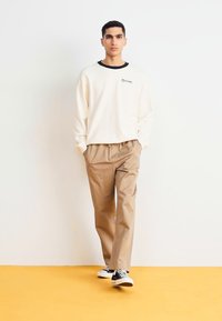 Krémová mikina s čiernym golierom, béžové nohavice a čierno-biele tenisky. Jednoduchý dizajn, bez vzorov, voľný strih, jednoliaty pozadie.