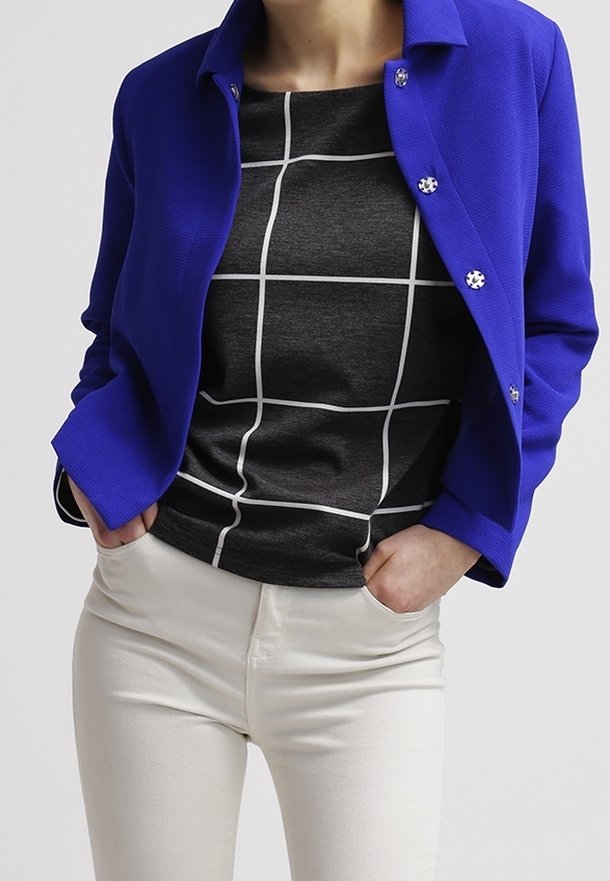Blazer blu con dettagli sui bottoni sopra una maglia a quadri bianco e nero, abbinato a pantaloni bianchi fitted. Tessuto liscio con design strutturato.
