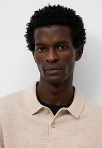 Polo shirt beige a maniche corte, texture morbida, con patta a due bottoni, indossato su una camicia scura. Lo sfondo è semplice e di colore chiaro.