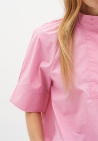 Kvinna med långt blont hår som bär en lös rosa kortärmad skjorta med hög krage mot en enfärgad vit bakgrund.