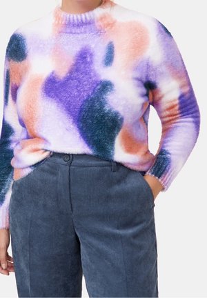 Personne portant un pull duveteux avec des motifs abstraits violet, rose et orange, associé à un pantalon en velours côtelé bleu marine taille haute.