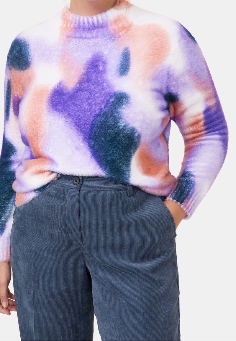 Personne portant un pull duveteux avec des motifs abstraits violet, rose et orange, associé à un pantalon en velours côtelé bleu marine taille haute.