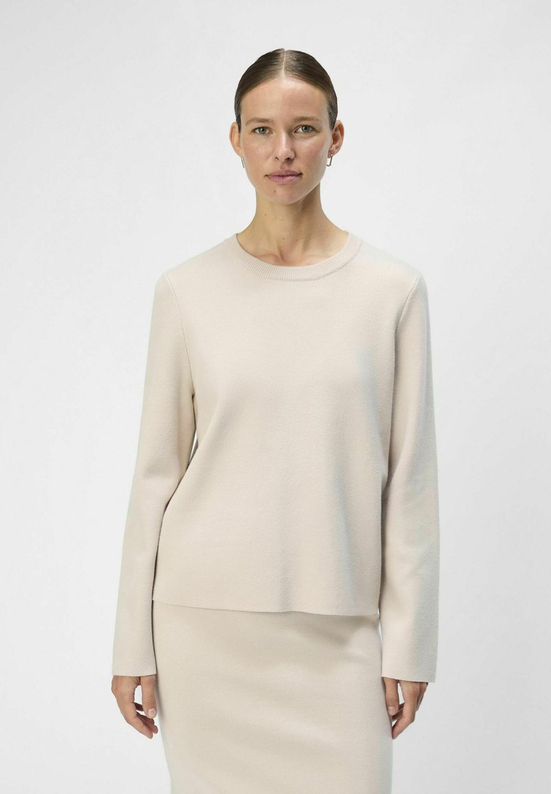 Ljusbeige tröja med långa ärmar och rundad halsringning, tillverkad av mjukt material. Bärs med en matchande kjol för en samordnad look.