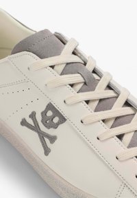 Zapatilla de cuero en color blanco roto con acentos de ante gris. Presenta un diseño de calavera y huesos cruzados en gris claro, cordones planos y detalles perforados.