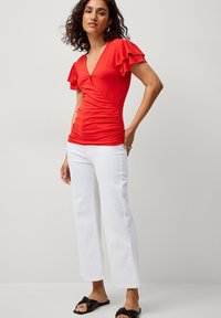 Rood aangesloten top met gerimpelde mouwen, gecombineerd met witte rechte broek. Model draagt zwarte slippers. Effen achtergrond.