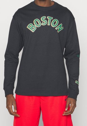 Person iført en sort langærmet trøje med "BOSTON" i grønne bogstaver og lyserøde atletikshorts.