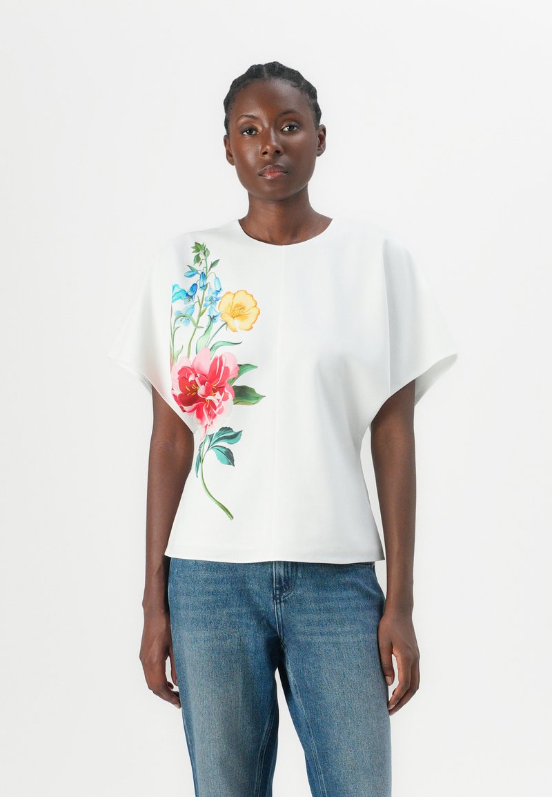 White short-sleeve top featuring colorful floral prints in pink, yellow, and blue. Soft texture with a relaxed fit, paired with denim.