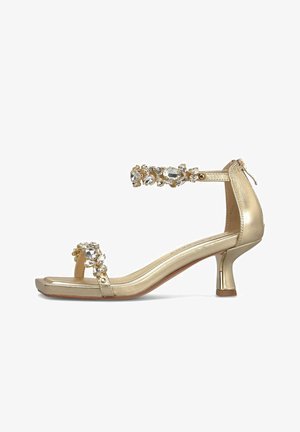 Złoty sandał z odkrytymi palcami, niskim zakrzywionym obcasem, paskiem wokół kostki i paskiem na palce zdobionym dużymi przezroczystymi kryształkami.