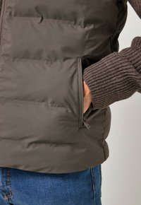 Chaleco de plumas marrón oscuro con tejido acolchado horizontal, bolsillo lateral con cremallera y manga de suéter de canalé visible por debajo. También se muestran unos vaqueros de denim.