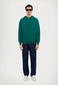 Grønn hettegenser i bomullsblanding med kengurulomme. Kombinert med mørkeblå jeans og hvite joggesko. Solbriller fullfører antrekket.