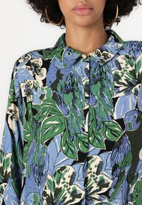 Floral and bird patterned blouse with a blue, green, black, and cream color scheme; features a collar and button detail.
