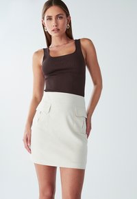 Ženska oblečena v brezrokavni rjav top in svetlo kremno mini krilo z velikima sprednjima žepoma, stoji ob navadni podlagi.