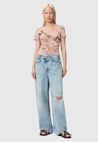 Femme aux longs cheveux blonds portant un haut rose à fleurs froncé, un jean bleu clair déchiré ample et des sandales noires, debout devant un fond uni.