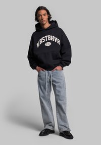 Felpa nera con scritta bianca "WRSTBHV" e tasca, abbinata a jeans baggy blu chiaro e scarpe nere, posizionata su uno sfondo semplice.