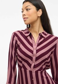 Rotes Hemd mit hellrosa horizontalen Streifen; verfügt über einen Kragen, lange Ärmel und eine taillierte Silhouette.