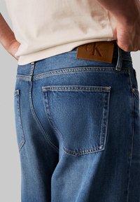 Jeans de denim azul con una cintura alta, que presentan un parche de cuero con el logo, costuras en contraste y cinco bolsillos. La textura es suave con un ligero brillo.