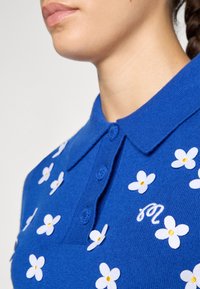 Blå polo skjorte laget av strikket stoff med hvit blomsterbroderi med gule midtpunkter, krage og placket med tre knapper.