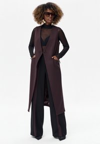 Długa kamizelka bez rękawów w ciemnym bordowym materiale z pojedynczym guzikiem, połączona z czarnym, przezroczystym topem i szerokimi czarnymi spodniami z paskami po bokach.