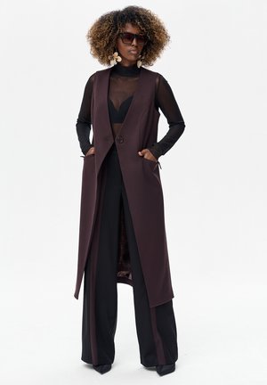Długa kamizelka bez rękawów w ciemnym bordowym materiale z pojedynczym guzikiem, połączona z czarnym, przezroczystym topem i szerokimi czarnymi spodniami z paskami po bokach.
