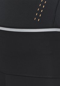 Zbliżenie czarnego materiału z przeszyciami, małymi brązowymi perforacjami i poziomym odblaskowym srebrnym paskiem jako detalem.