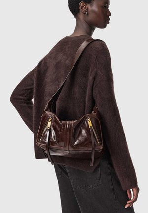 Mujer con suéter marrón oscuro de peluche y jeans negros, llevando un bolso de hombro de cuero marrón oscuro con cremalleras doradas en la espalda.