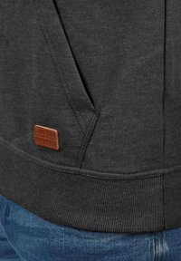 Sudadera gris oscuro con una textura suave, con un bolsillo tipo canguro y una etiqueta de cuero marrón. El dobladillo y los puños acanalados añaden detalle.