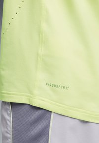 Jasnożółta sportowa koszulka wykonana z lekkiego materiału, cechująca się gładką fakturą, widocznym przeszyciem oraz zielonym logo „CLOUDSPUN”.