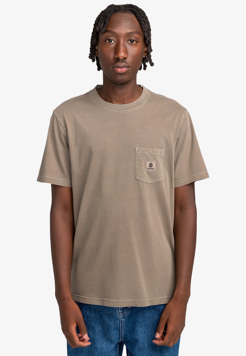 T-shirt z krótkim rękawem i okrągłym dekoltem w stonowanym brązowym kolorze. Posiada kieszonkę na piersi z naszywką z logo. Noszony z niebieskimi dżinsami.