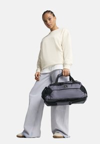 Bolsa de deporte gris con acentos negros y paneles de malla. Cuenta con asas dobles, una correa ajustable y un parche con el logotipo en la parte frontal.
