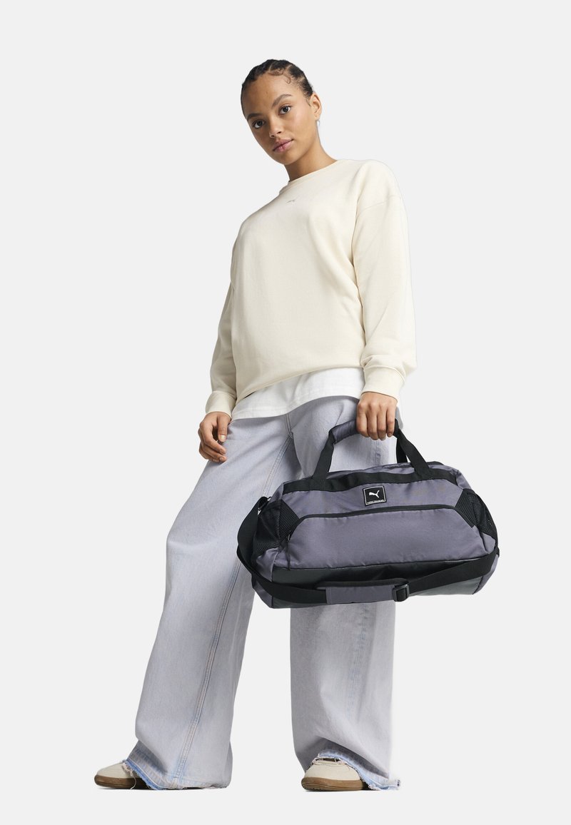 Bolsa de deporte gris con acentos negros y paneles de malla. Cuenta con asas dobles, una correa ajustable y un parche con el logotipo en la parte frontal.