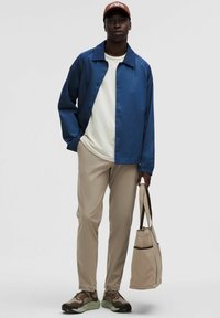 Lagana plava jakna preko bijele košulje, bež hlače, smeđitenisice s zelenim detaljima i bež torba, nosi osoba koja stoji protiv sive pozadine.